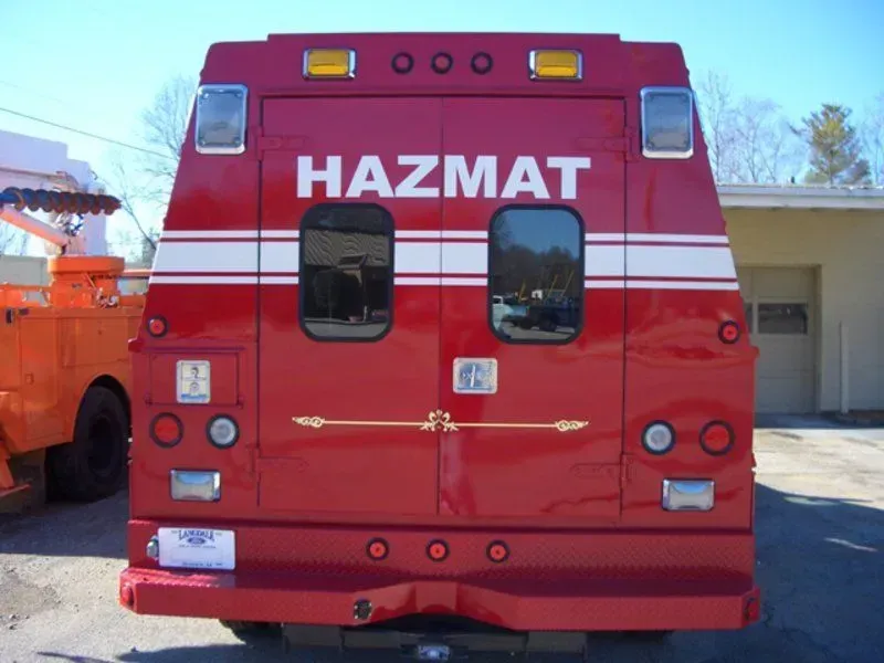Rear of a red HAZMAT truck with white stripes, windows, and lights, parked outdoors.