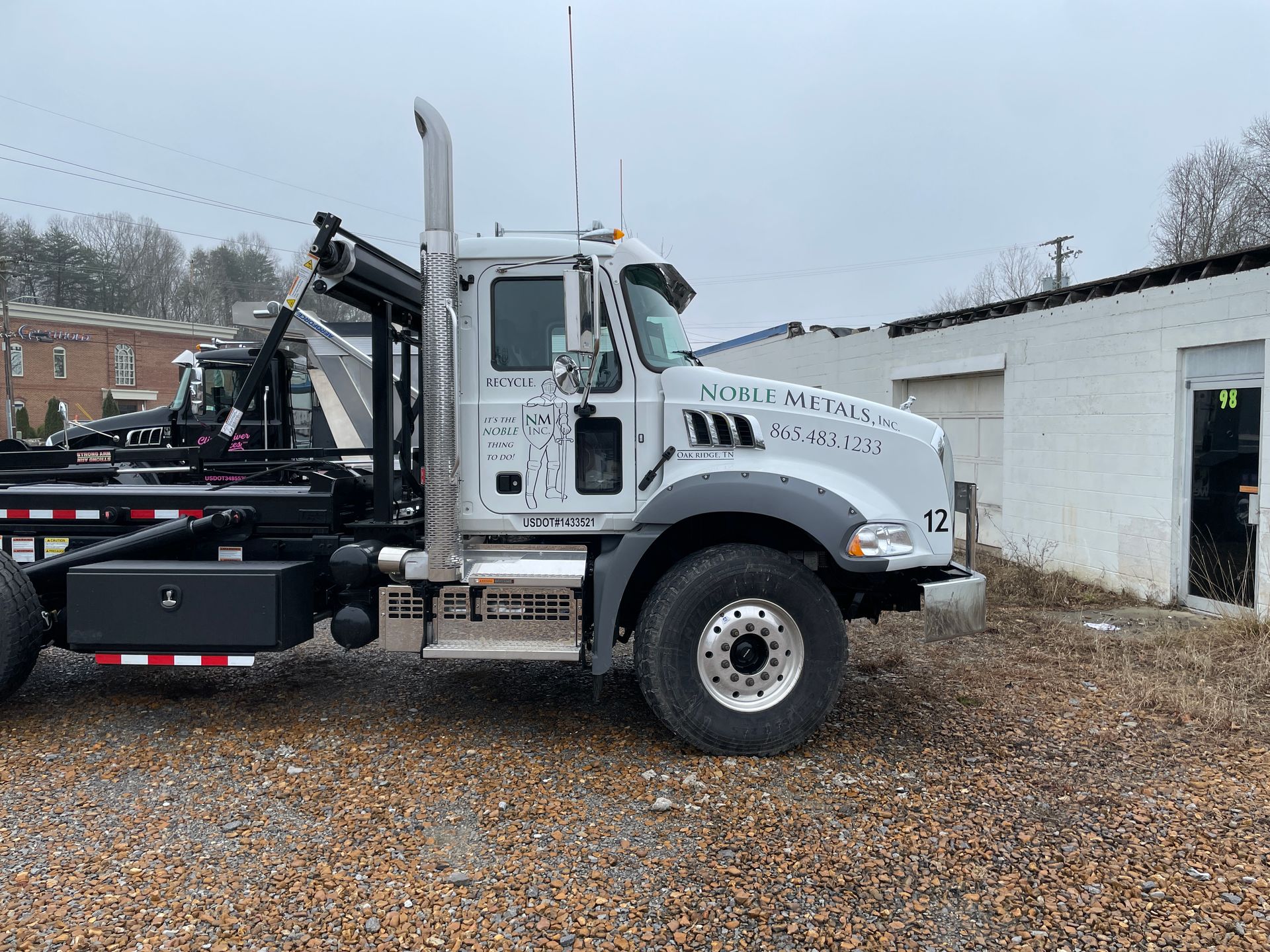 White Mack roll-off truck parked by a white building on a gravel lot.