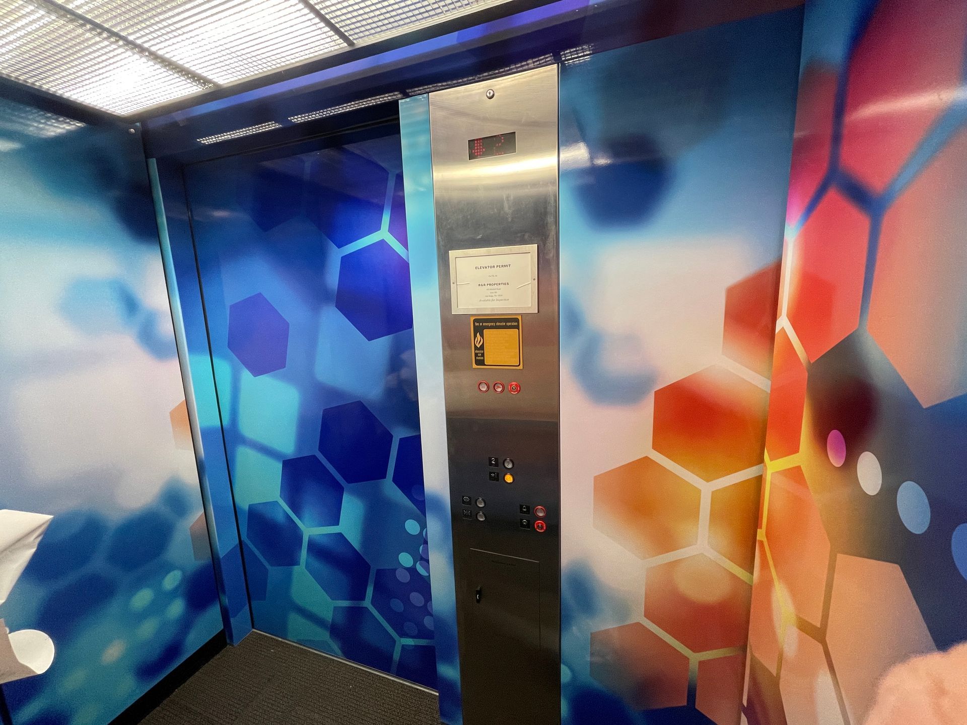 Elevator interior with colorful geometric design. Stainless steel control panel, doors open.