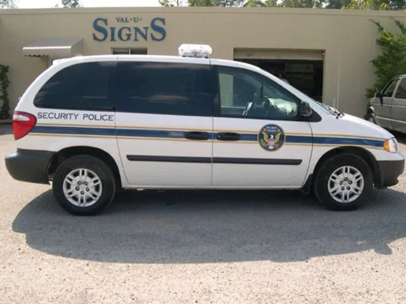 White security police van with blue stripe and emblem, parked in front of a sign shop.