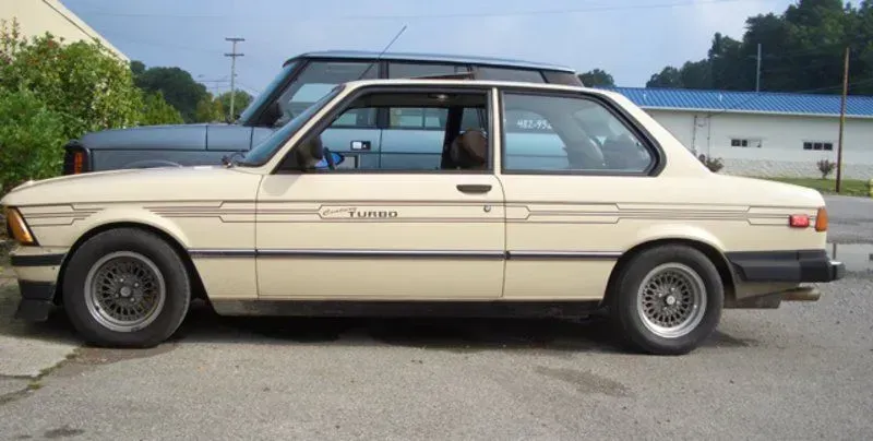 Beige vintage BMW coupe with turbo badge, parked on asphalt, with a gray SUV in the background.
