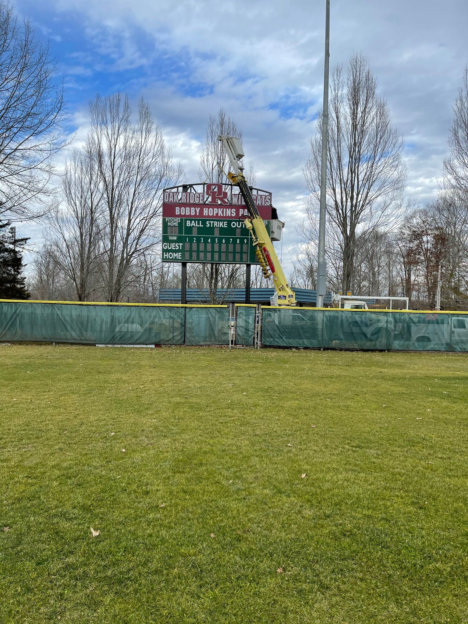 Baseball scoreboard with a lift being used for maintenance. Green fence, grass, and trees are in the foreground.
