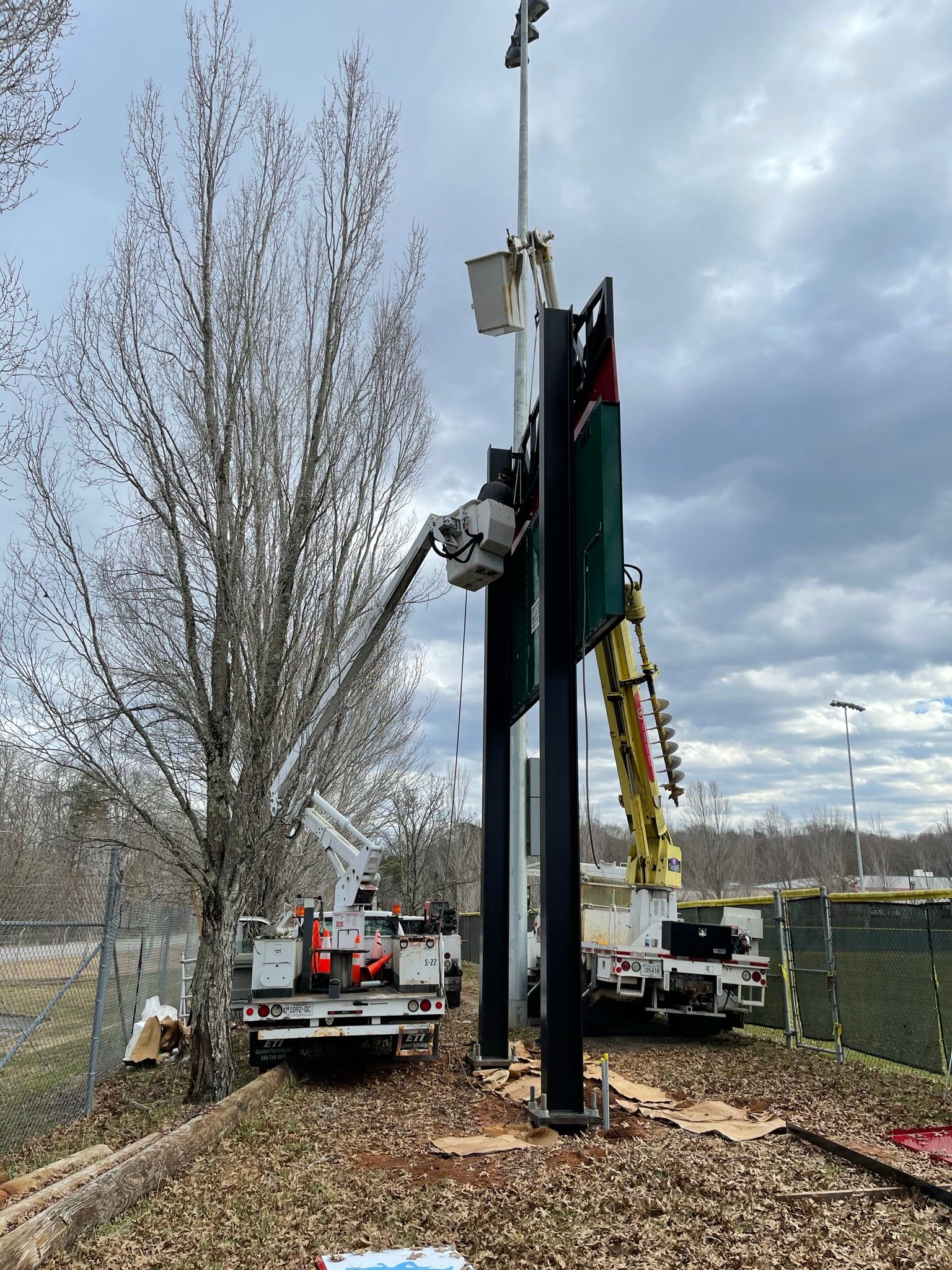 Two utility trucks near a tall, black sign with a traffic signal on a cloudy day. Workers in the lifts.