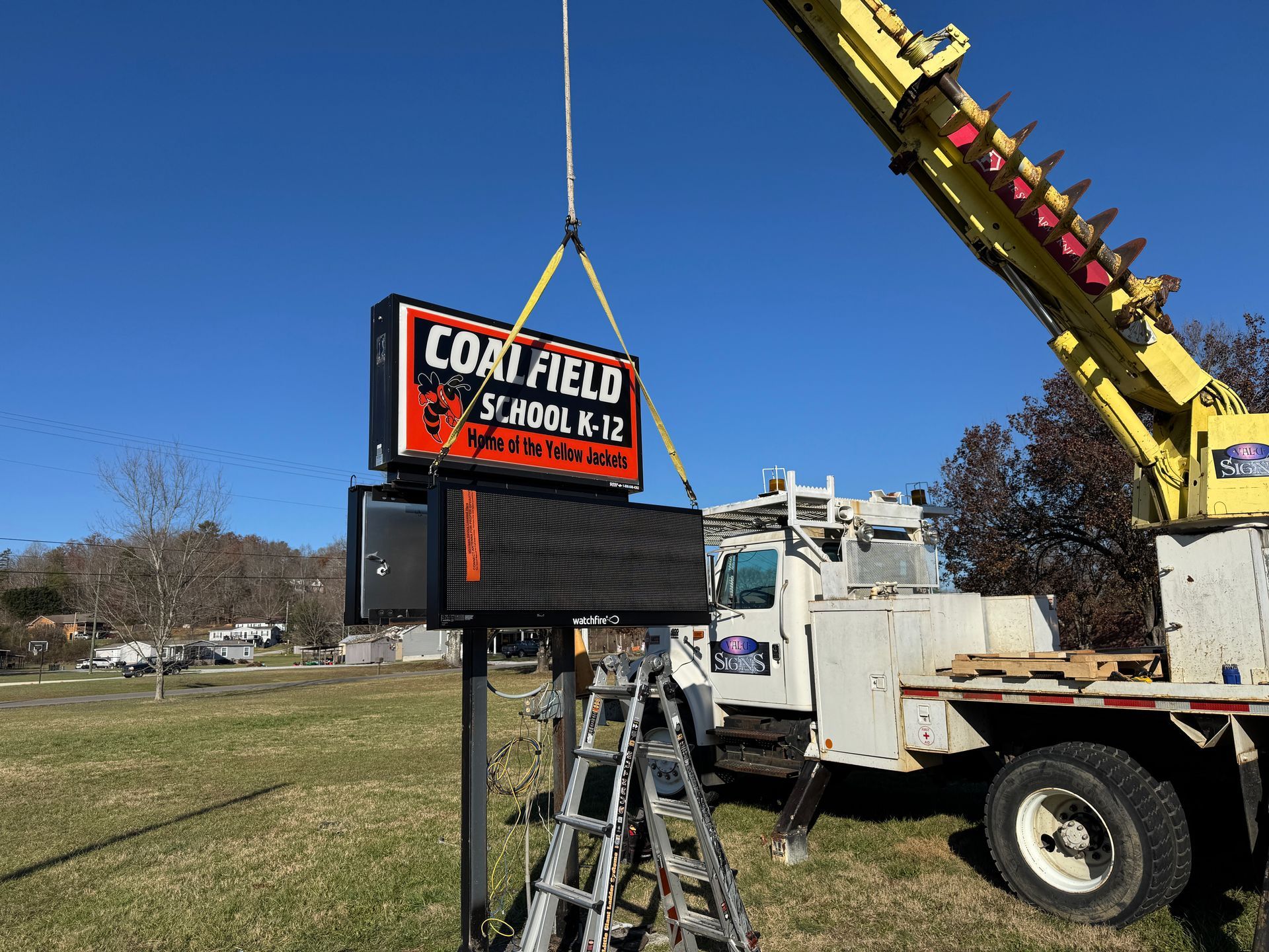 A crane lifts a new sign for Coal Field School K-12, atop a pole, near a truck, on a grassy area under a blue sky.