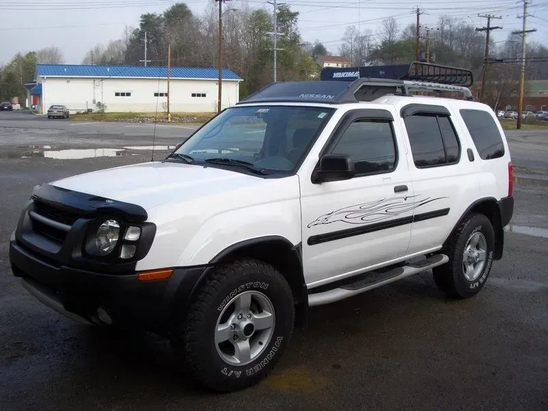 White Nissan Xterra SUV with black accents, roof rack, and side steps parked on a paved lot.