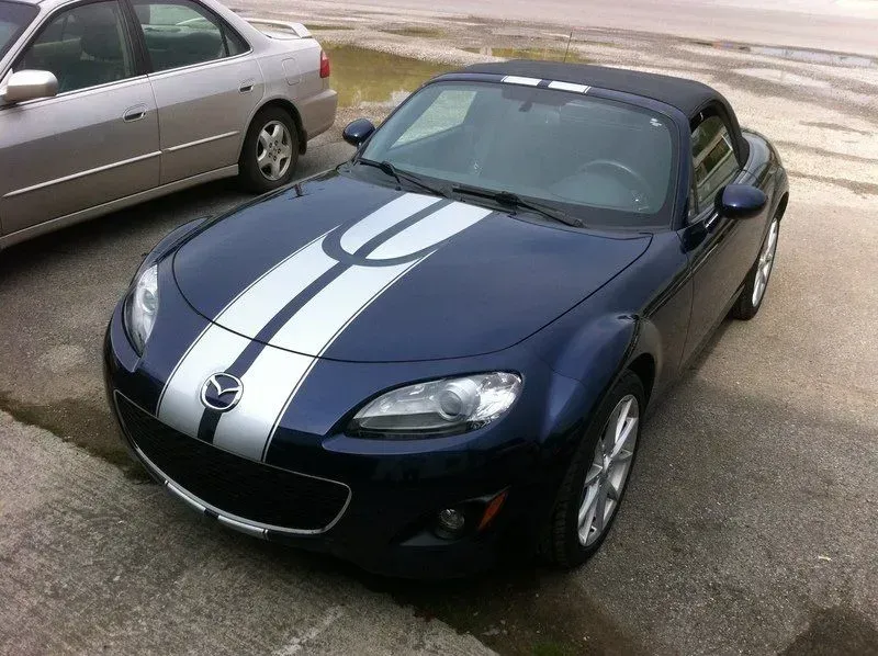 Blue Mazda Miata convertible with silver racing stripes parked near a silver sedan on a concrete surface.