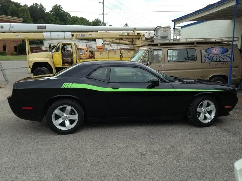 Black Dodge Challenger with green side stripe parked on a street near a van and a yellow truck.