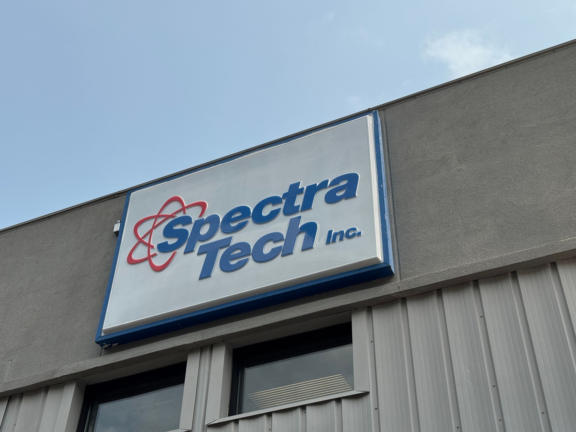 Spectra Tech Inc. sign on a gray building with a blue sky background. The sign has a blue border and logo.