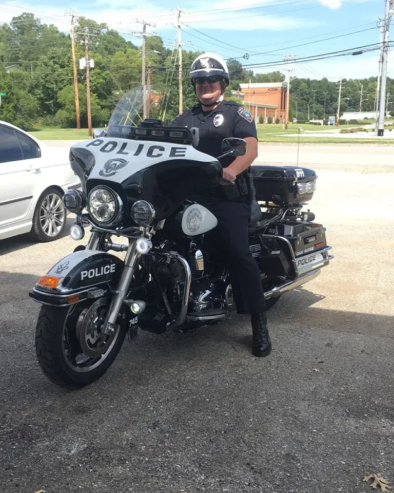 Police officer seated on a black motorcycle, wearing a helmet and uniform, outdoors.