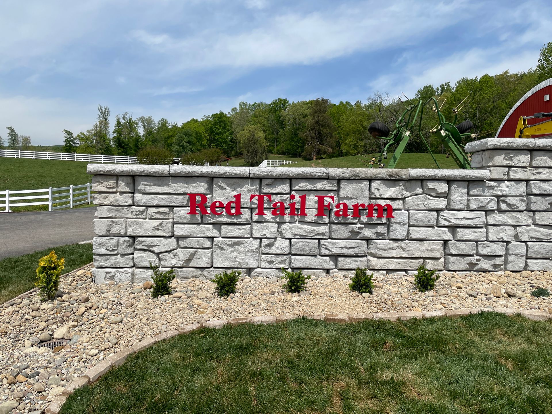 Red Tail Farm sign on a stone wall, red letters, against a field with white fences, and green grass.
