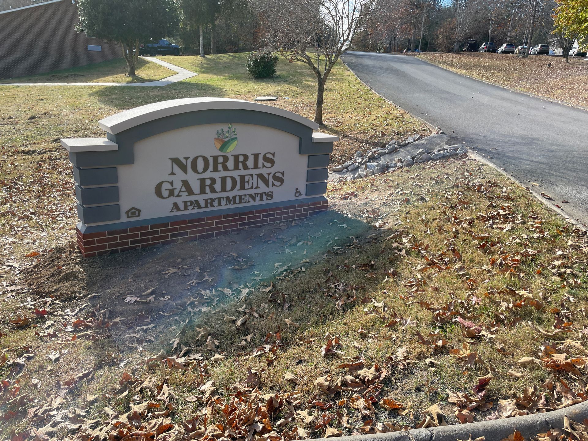 Sign for Norris Gardens Apartments; beige, brick, and gray with a road in background.