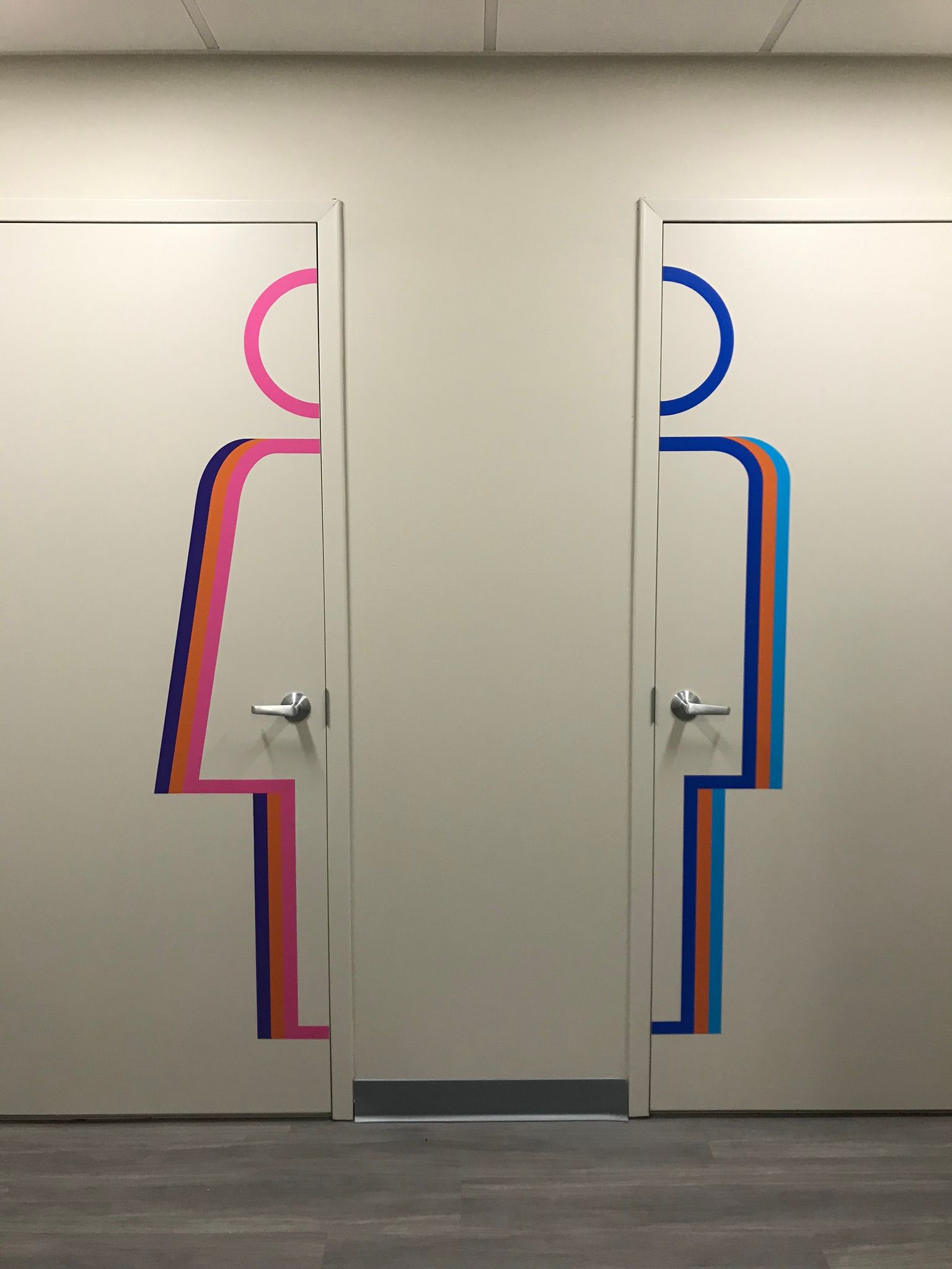 Two restroom doors with gender symbols: pink/purple woman and blue/teal man. Gray floor.