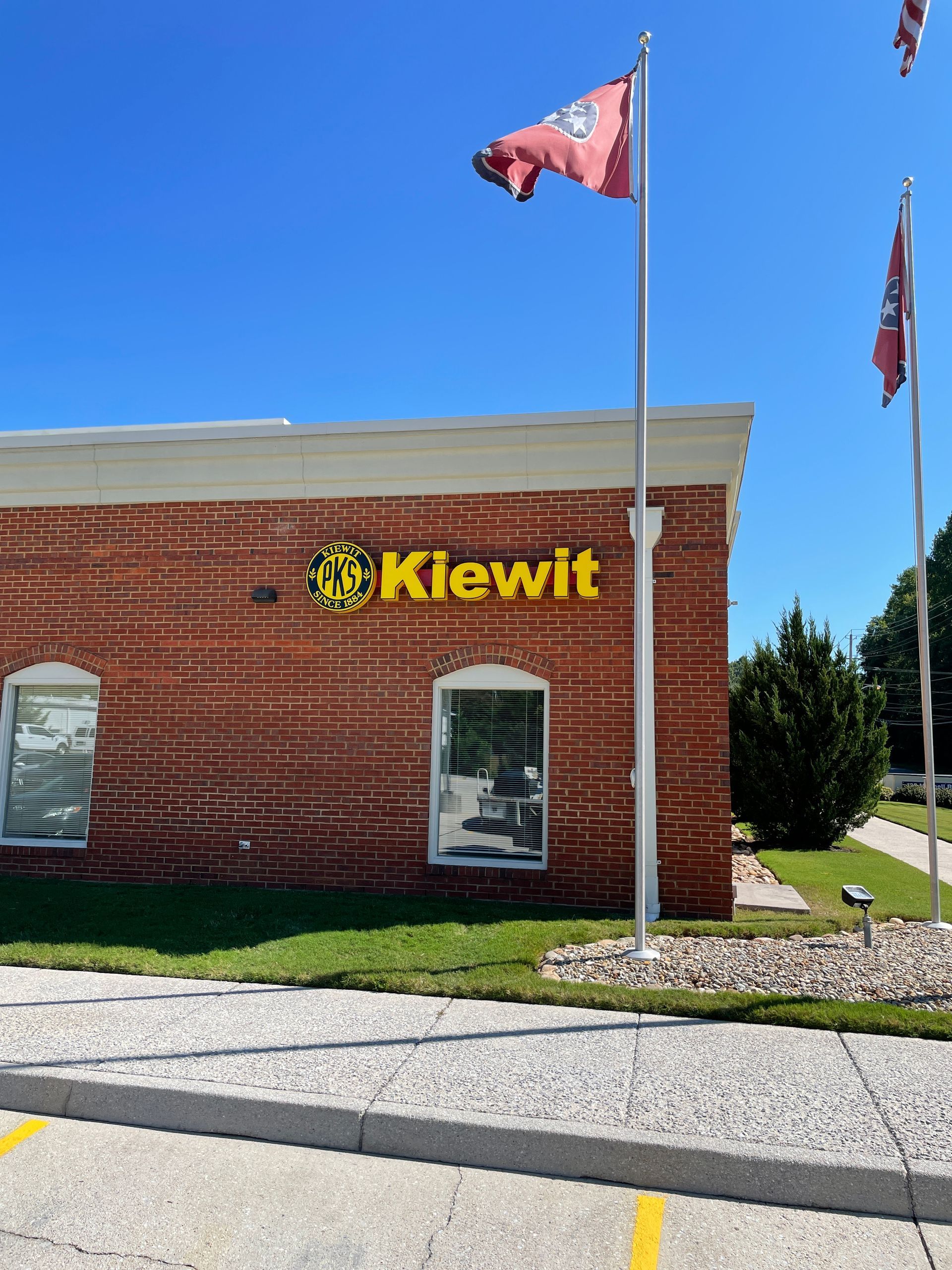 Kiewit building with yellow logo on brick exterior, flags in background, clear blue sky.