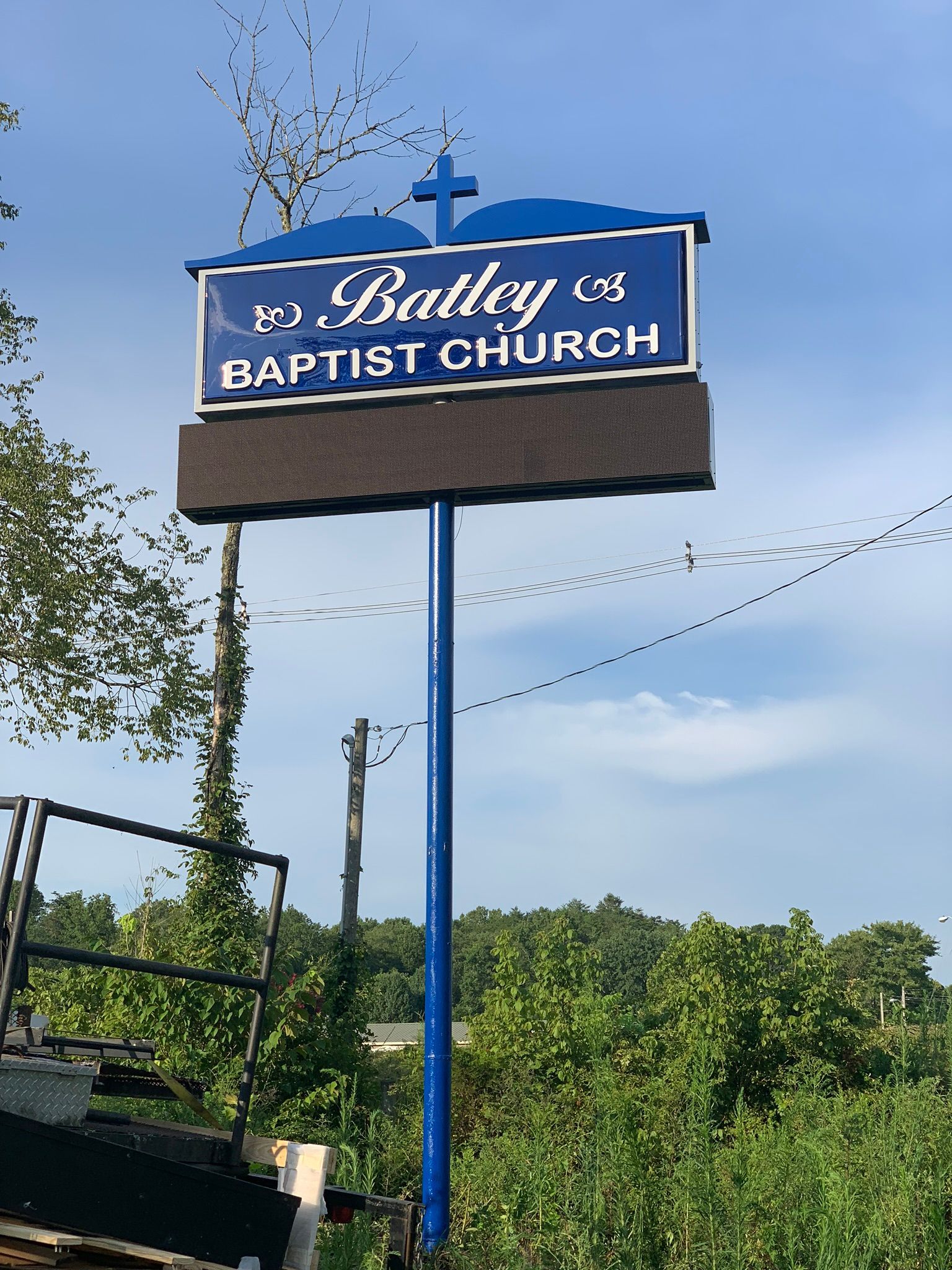 Sign for Bailey Baptist Church: blue with white lettering, cross at top, mounted on a pole, trees in background.