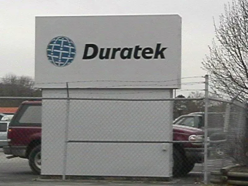 Duratek sign with logo above a white box structure, behind a chain-link fence, parked cars in background.
