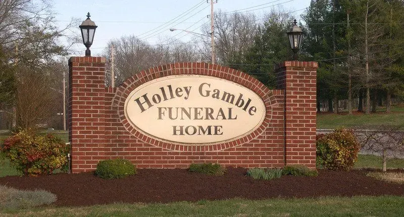 Sign for Holley Gamble Funeral Home, brick structure with black lamps, brown mulch, and green shrubs.