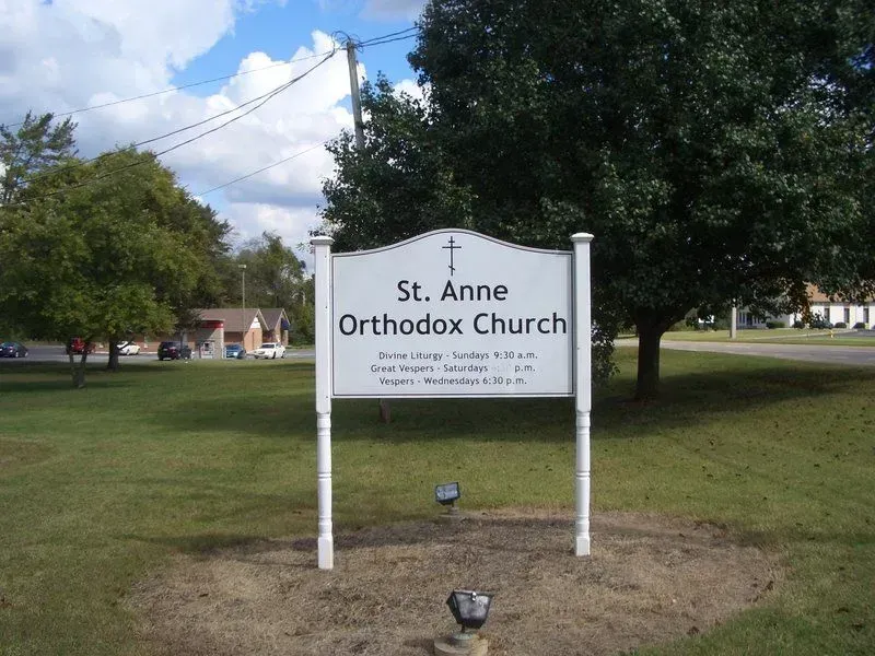 Sign for St. Anne Orthodox Church on a grassy lawn with a tree and a road in the background.