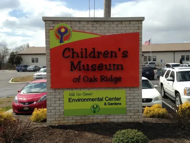 Sign for Children's Museum of Oak Ridge, orange and red with logo. Buildings and cars in background.