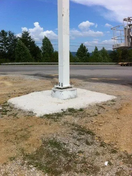 White flagpole base on a concrete square with gravel surroundings, road and trees in the background.