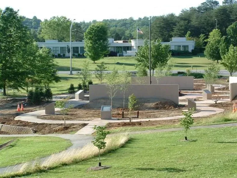 Park with a memorial feature, light tan walls and walkways, trees, green grass, and buildings in the background.