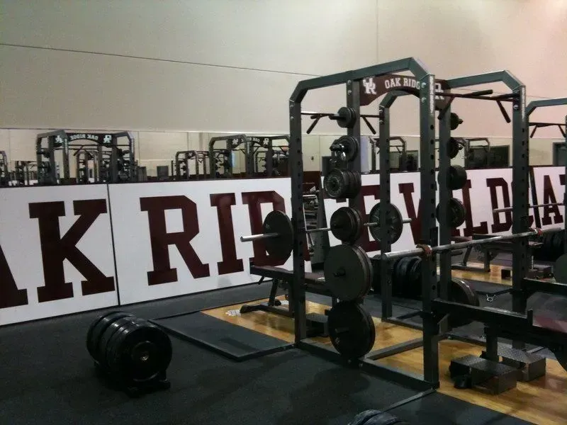 Weightlifting racks with barbells and plates in a gym; the wall displays 