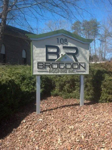 Sign for Brogdon Roofing Inc. at 108, with green and silver accents, set in front of shrubbery.