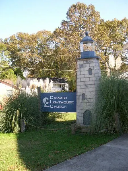 Calvary Lighthouse Church sign with a lighthouse sculpture, surrounded by bushes and trees.