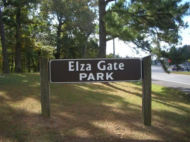 Elza Gate Park sign in a grassy area with trees, road visible in the background.