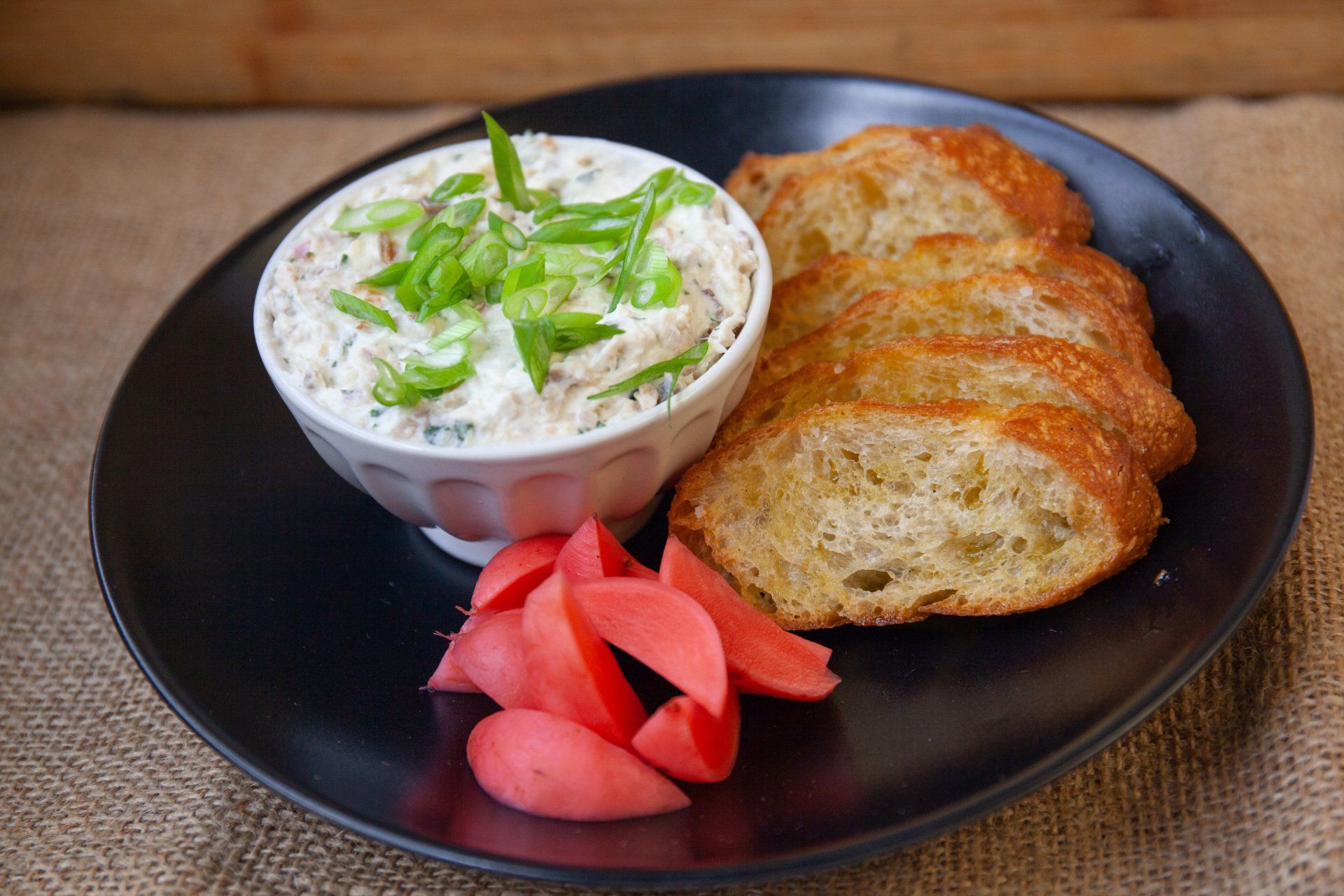 A black plate topped with a bowl of dip , bread and radishes.