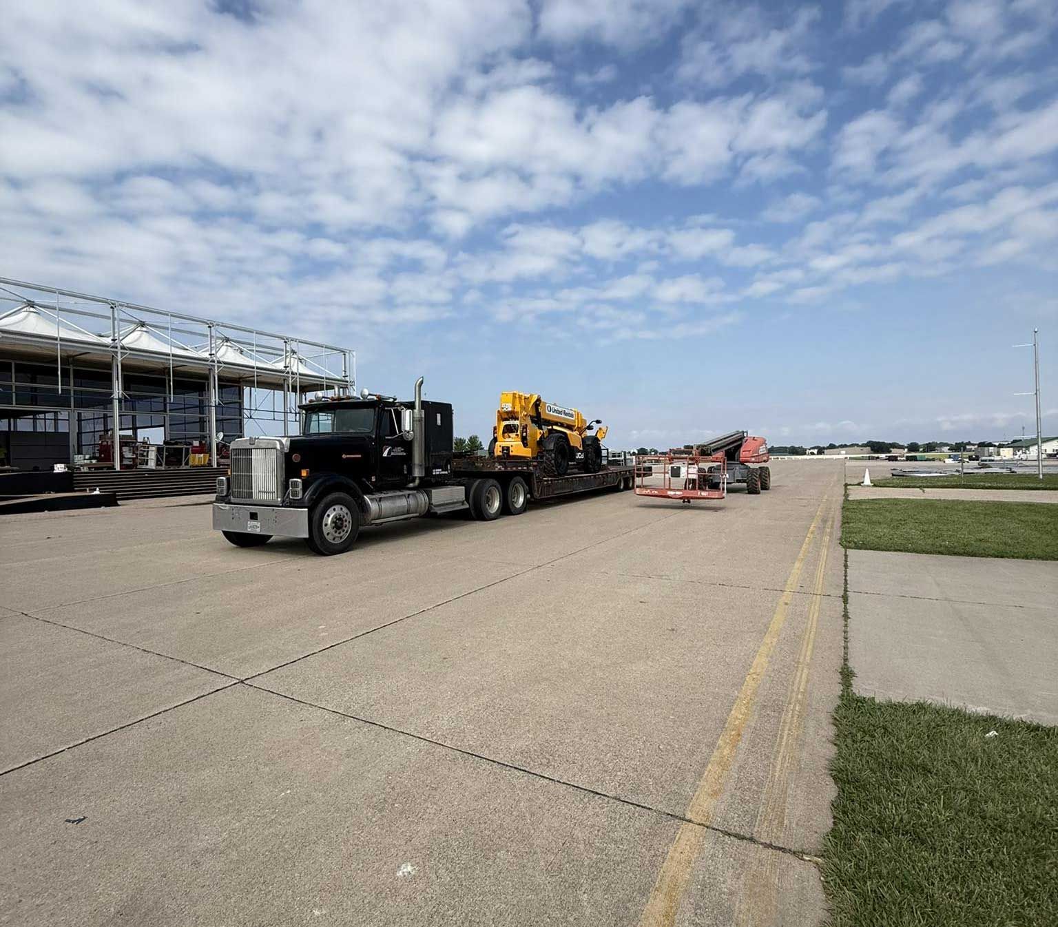 Tow truck hauling heavy equipment on a paved tarmac near an airport hangar under a cloudy sky
