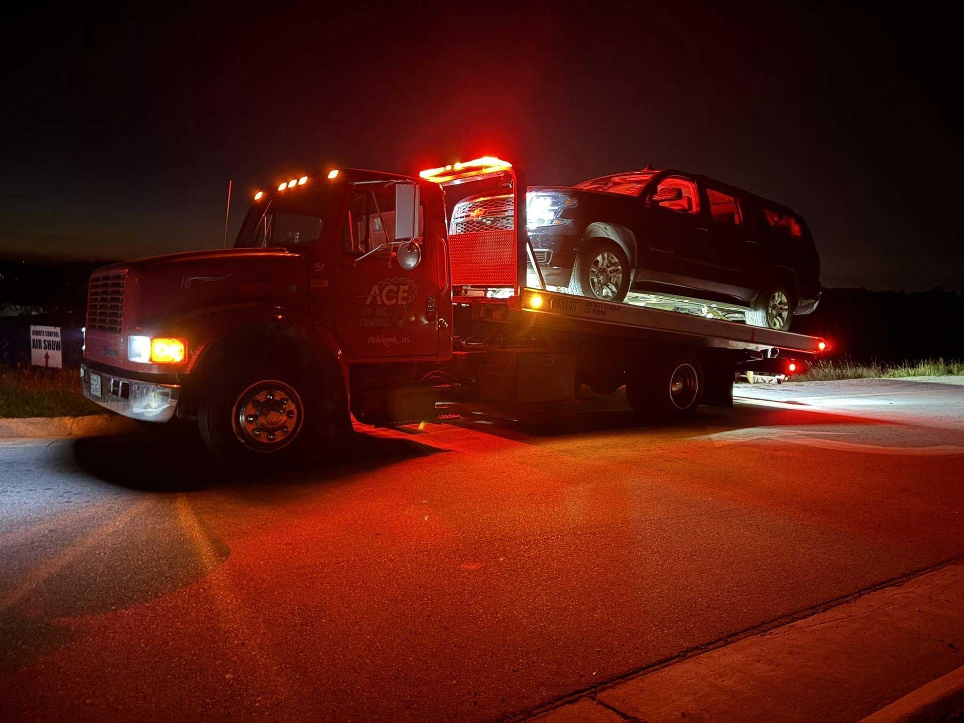 Tow truck hauling a dark SUV at night, lit by red and orange emergency lights.