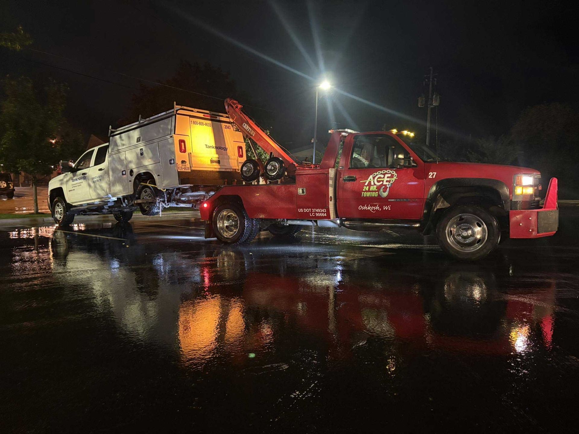 Red tow truck hauling a white vehicle at night on a wet, reflective street