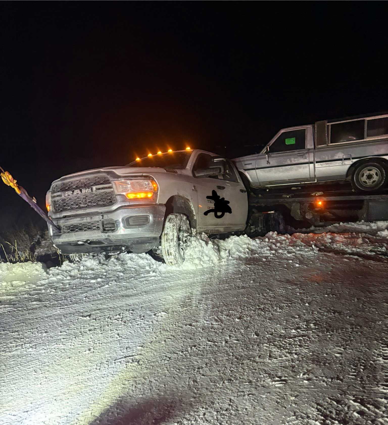 Pickup truck stuck in snowy ditch at night, with another truck on a tow bed nearby