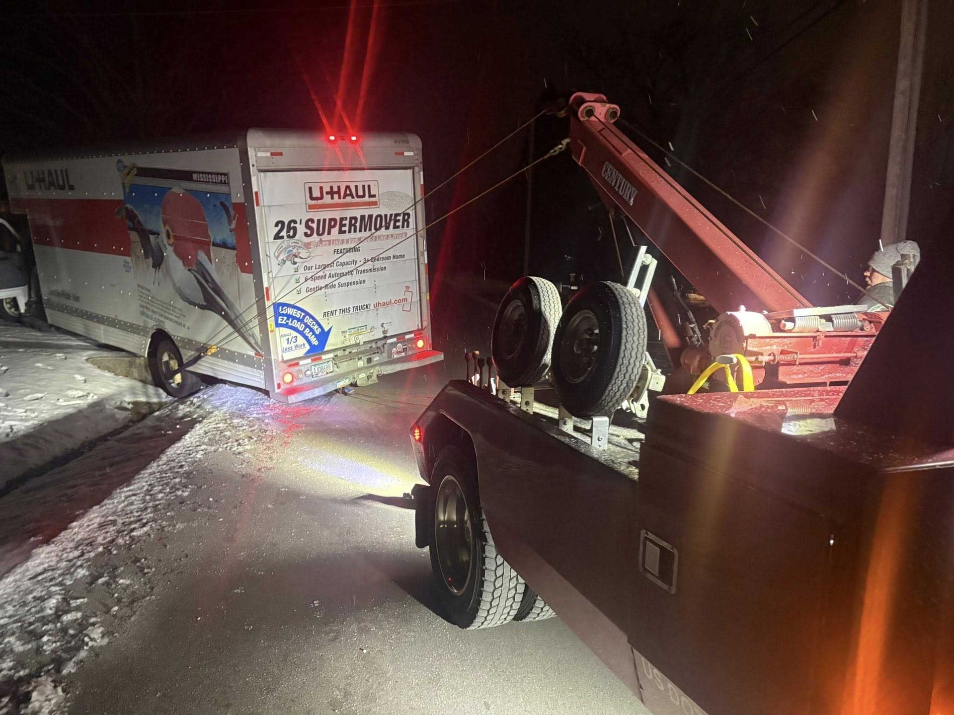 Tow truck recovering a white truck from a snowy roadside at night, with emergency lights visible