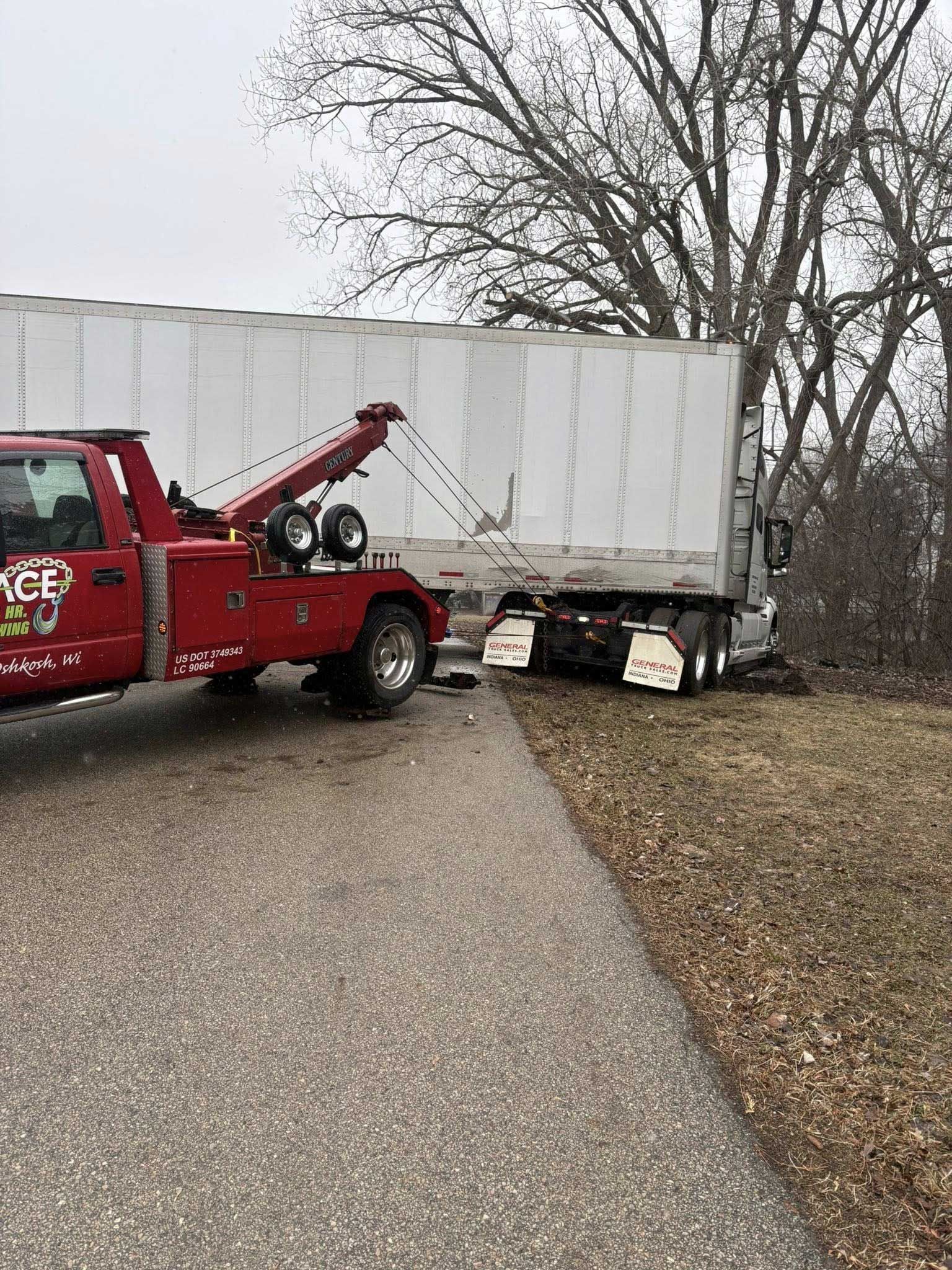 Red tow truck pulling a white trailer off a gravel road beside leafless trees.