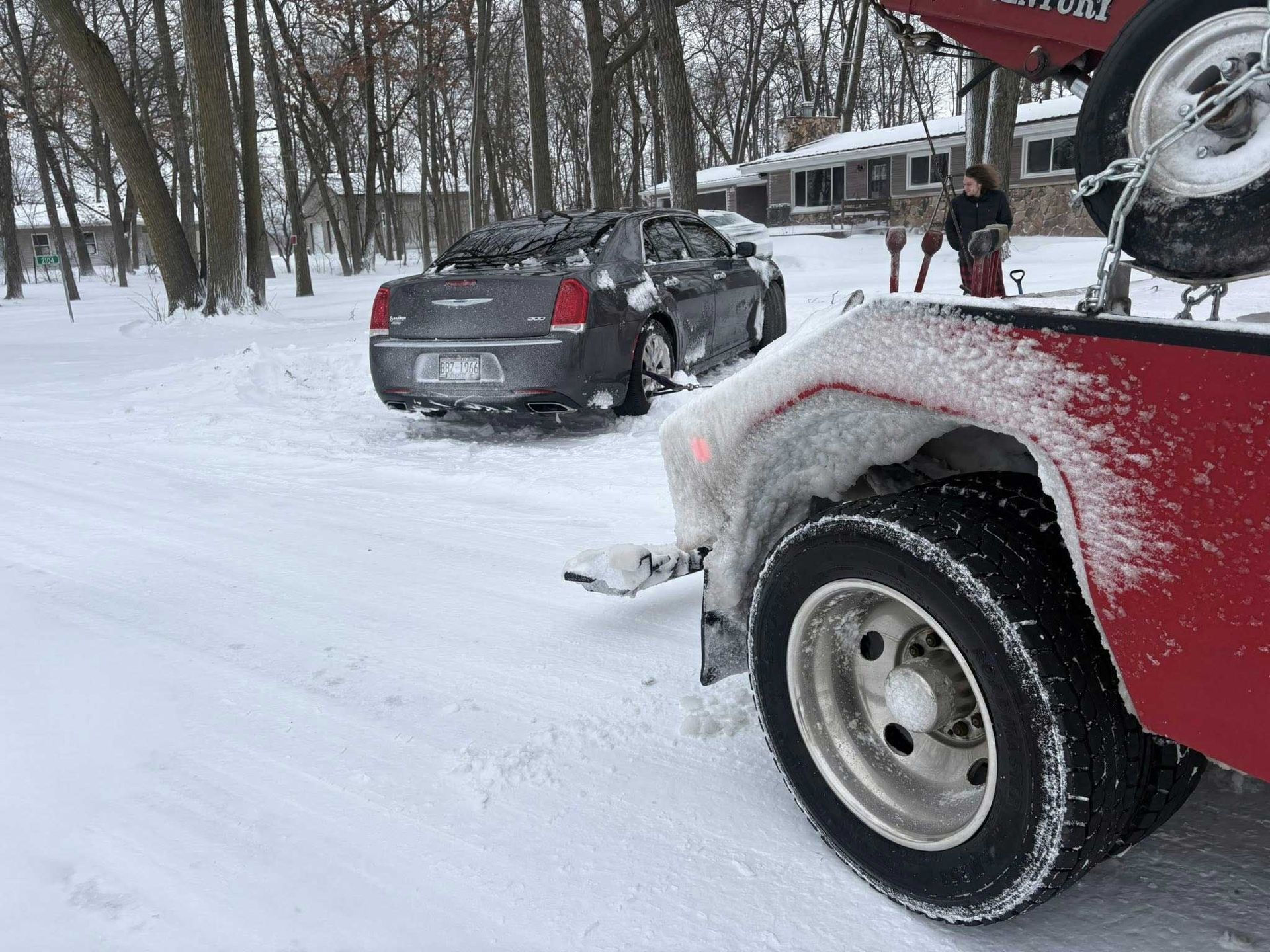 Snowy roadside scene with a gray car being towed by a red tow truck near a house.