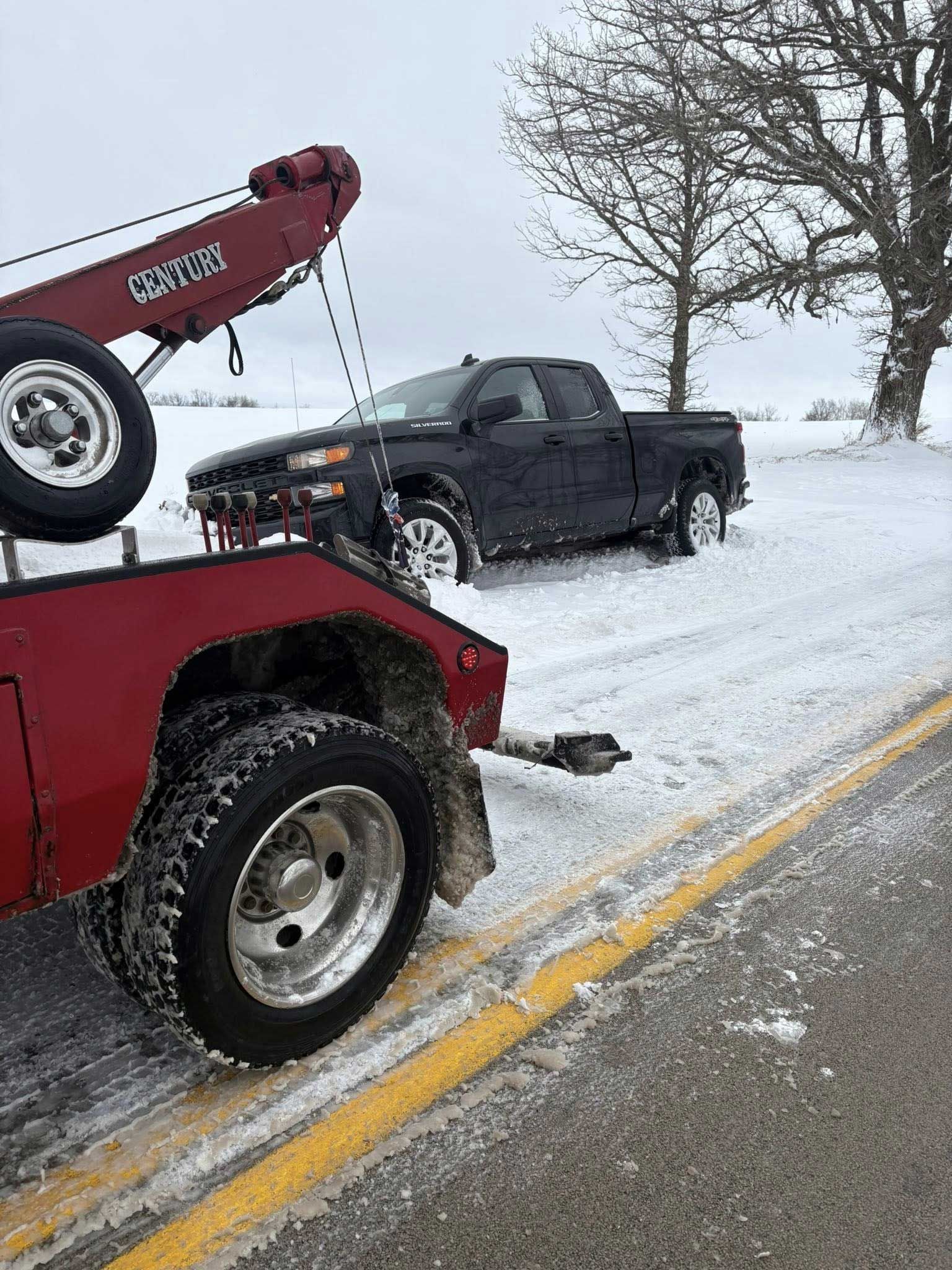 Tow truck towing a black pickup on a snowy roadside beside a tree