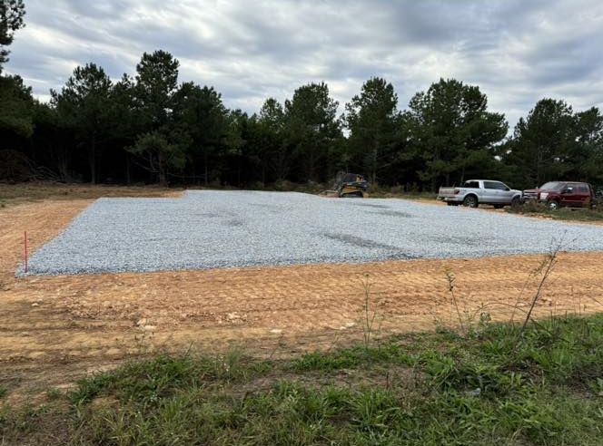 Gravel-covered foundation site, bordered by dirt, with a truck and trees in the background under a cloudy sky.