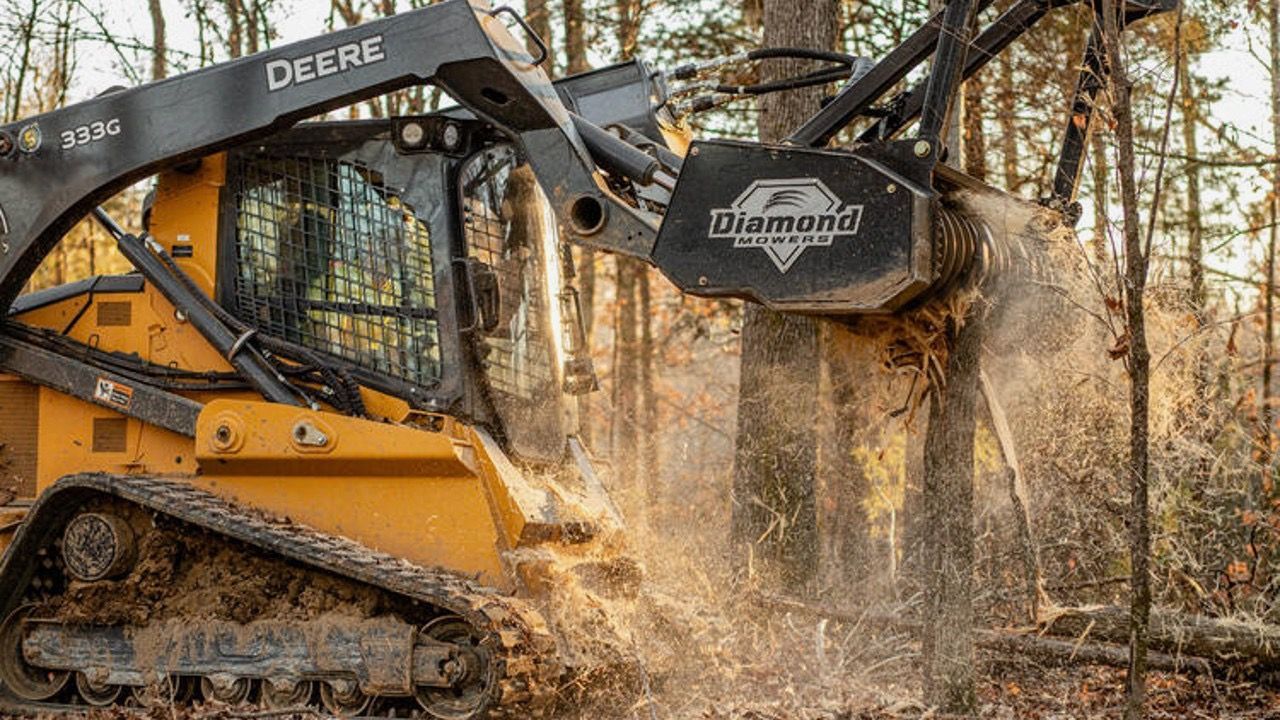 A yellow John Deere skid steer with a mulching head is cutting down a tree in a forest.