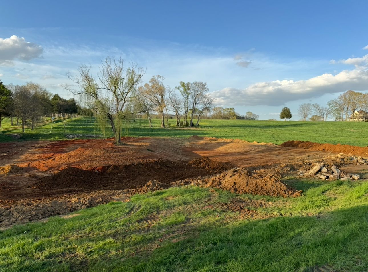 Dirt piles surrounded by green grass and trees under a cloudy blue sky.