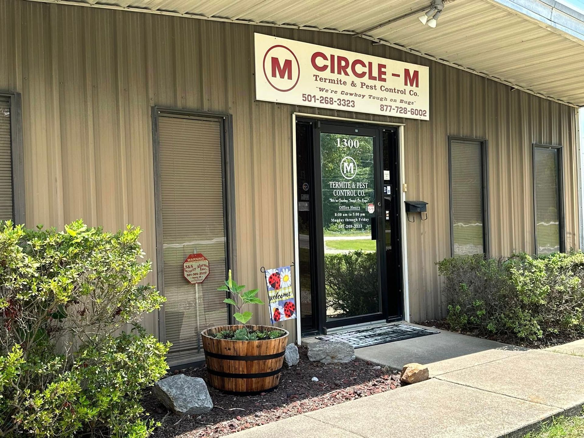 Exterior of Circle M business with sign above door. Wooden planter and bushes in front. Brown siding and a white trim.