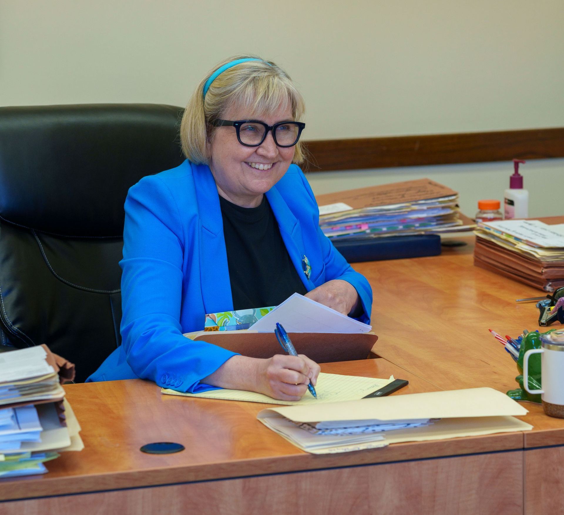 A woman in a blue jacket and glasses is sitting at a desk