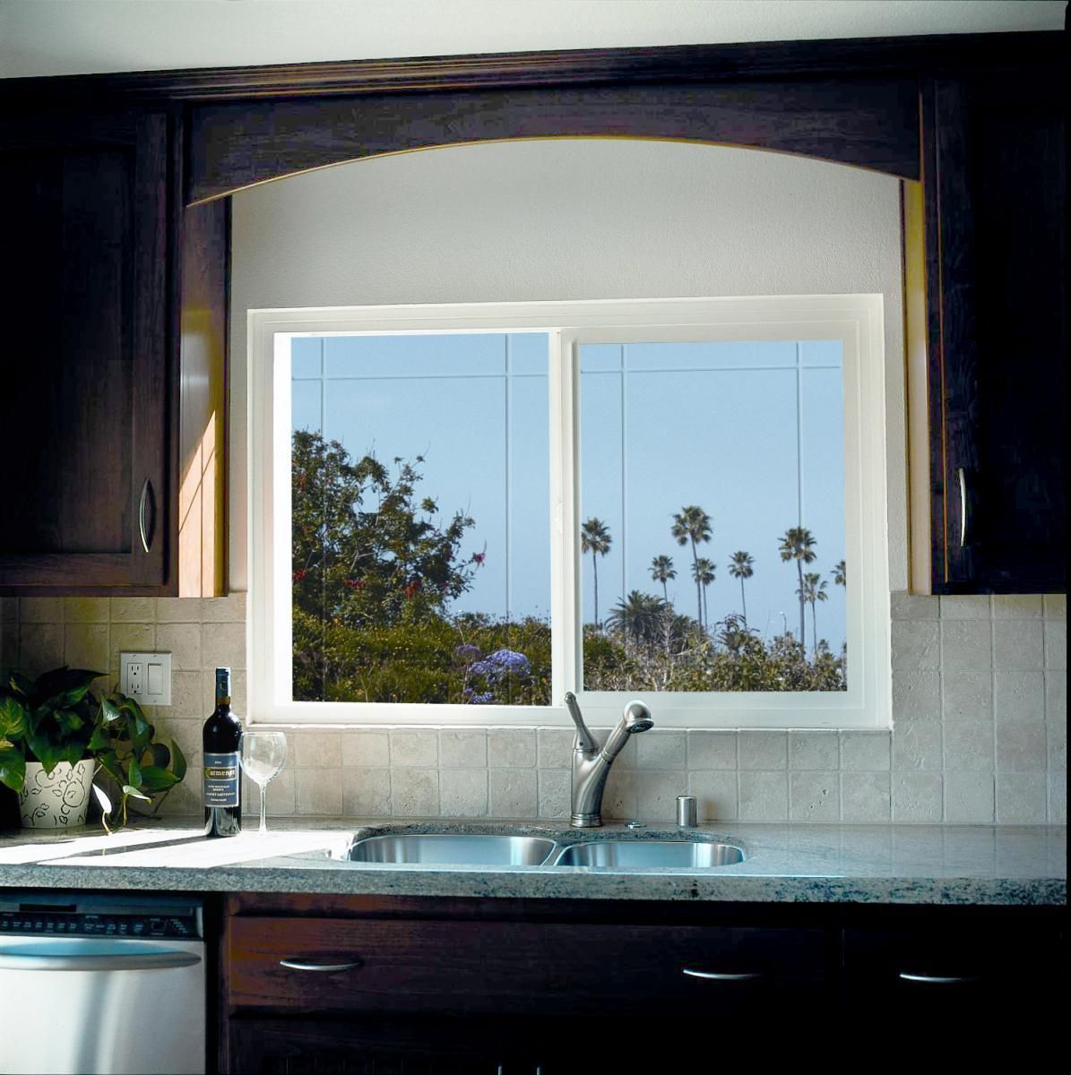 Kitchen sink beneath a window with a view of trees and palm trees, dark cabinets and white backsplash