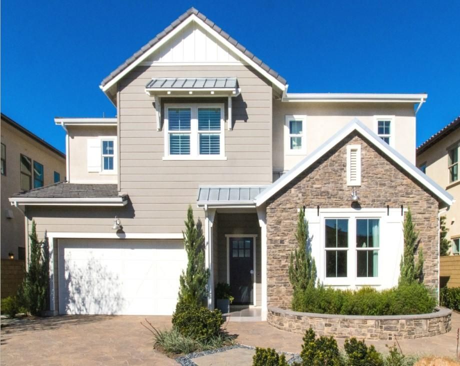 Two-story beige house with stone accents, white garage door, and front porch under a bright blue sky
