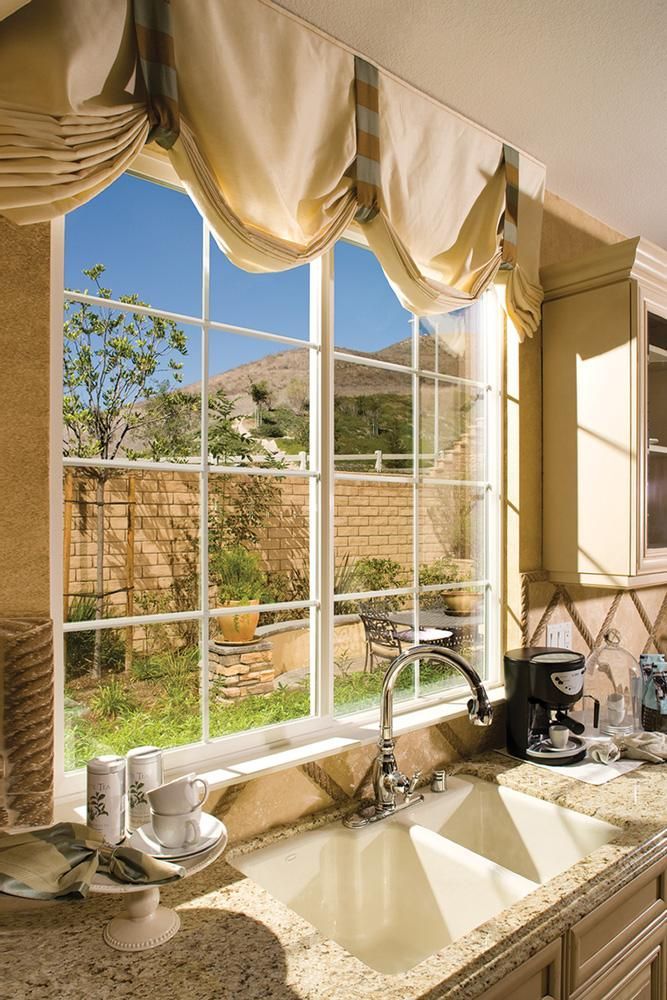 Sunlit kitchen sink under a large window with cream curtains and a view of the garden outside