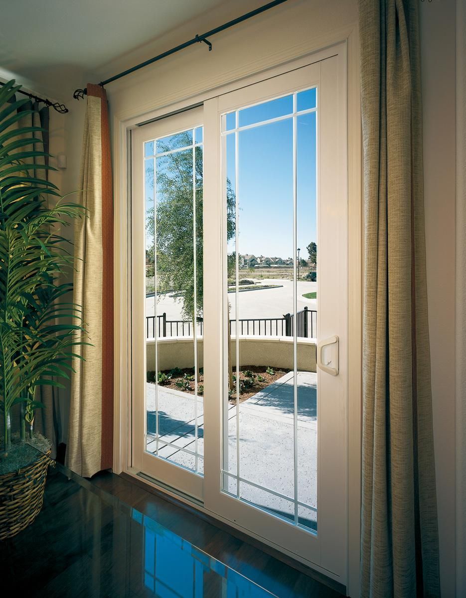 Sliding glass door with beige curtains opening to a sunny patio and blue sky outside