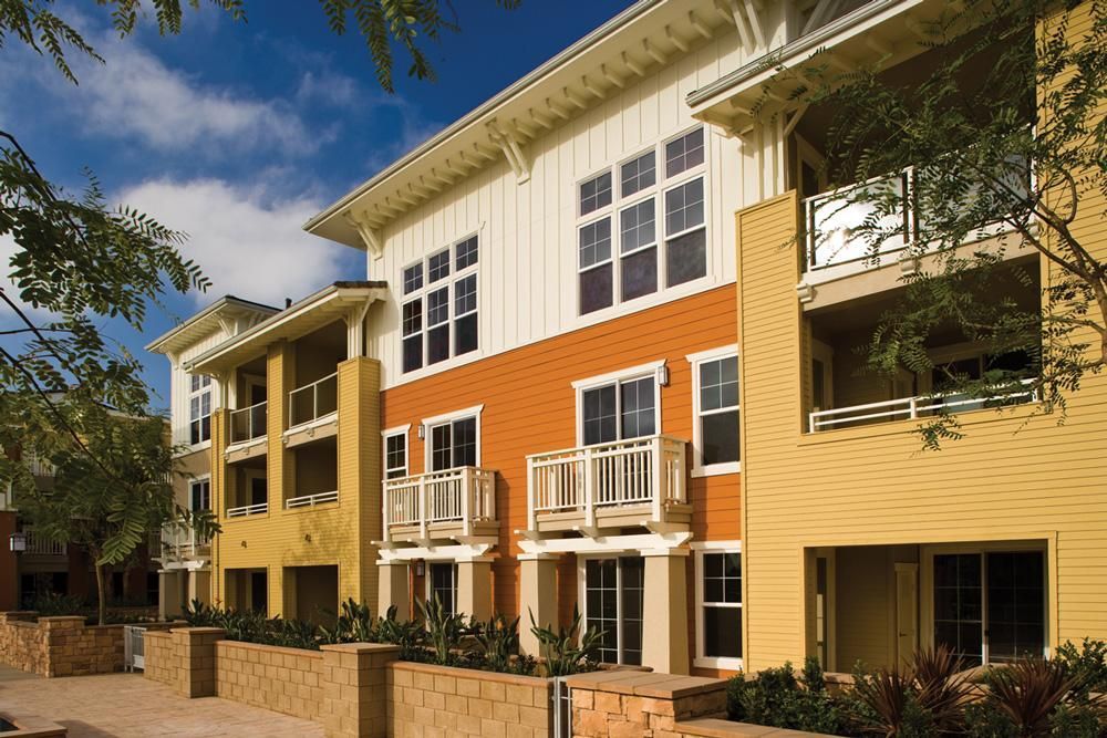 Modern apartment building with beige and orange exterior, balconies, and a landscaped front entrance under blue sky