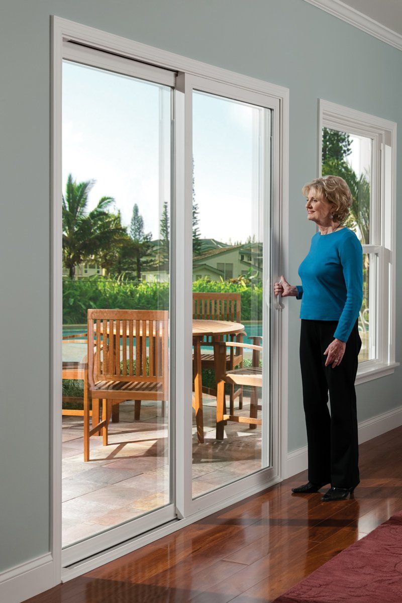Woman standing by a large sliding glass door, looking out at a patio with wooden chairs and a table.