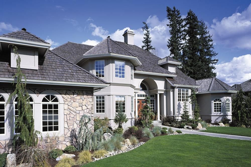 Large stone house with gray roof, arched entry, white trim, and manicured lawn under a blue sky