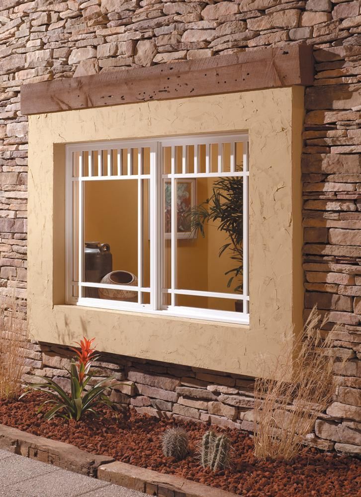 Stone house window with white bars and beige trim set in a rough stone wall.